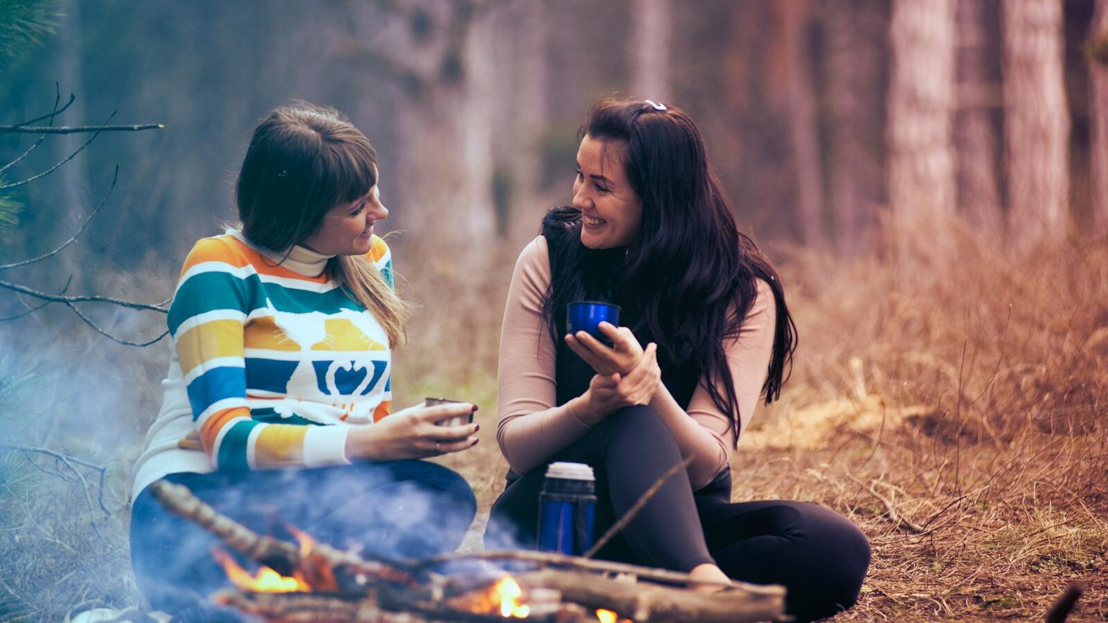 Two women enjoy coffee by a campfire in a forest, embracing the warmth and togetherness of nature. Depicting active listening in coaching